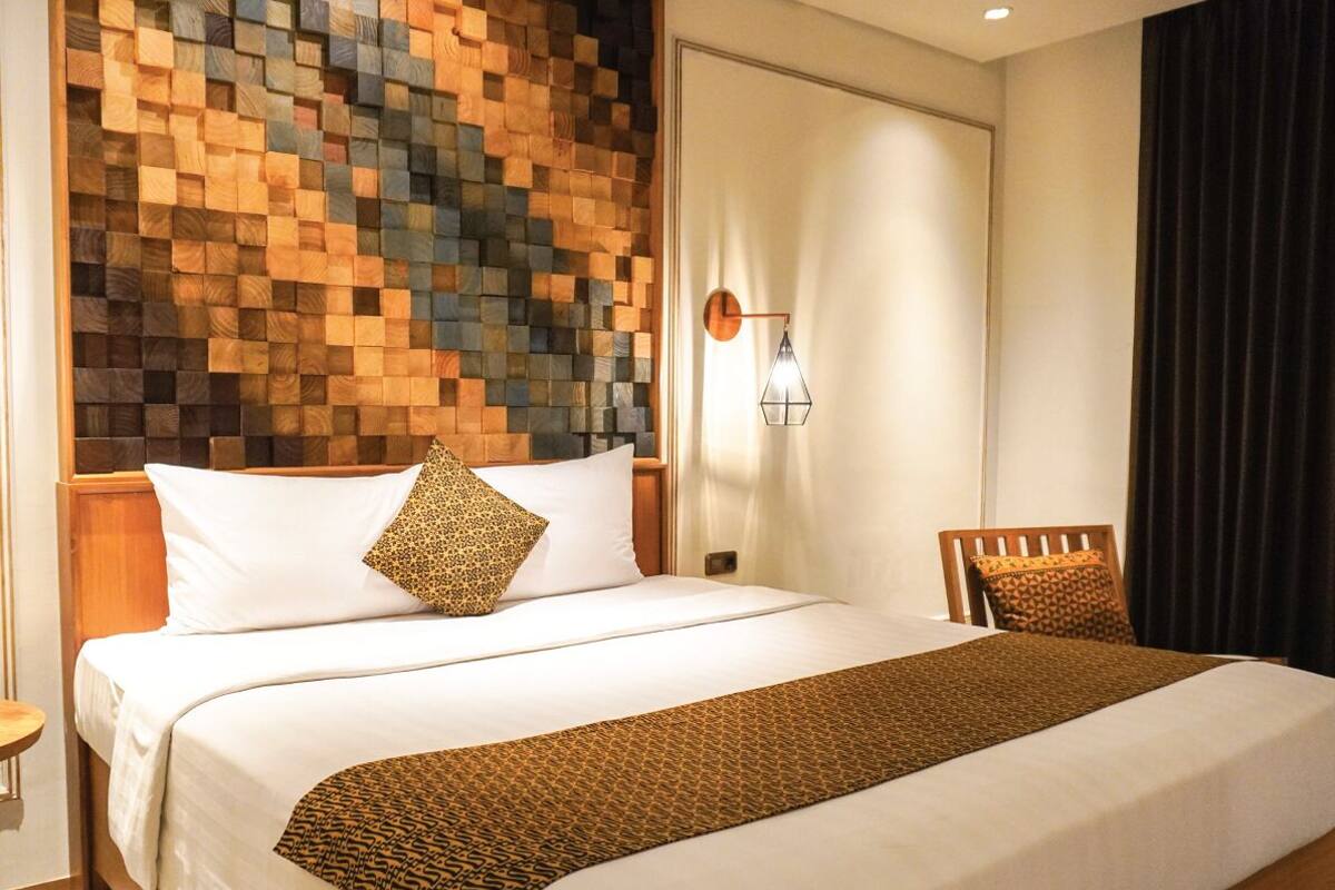 A hotel room with a large bed, a wooden headboard, a nightstand, a chair, and a wall-mounted lamp.