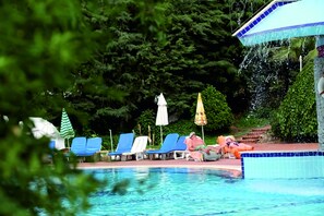 Zona de piscina con sillas de descanso y sombrillas, rodeada de vegetación.