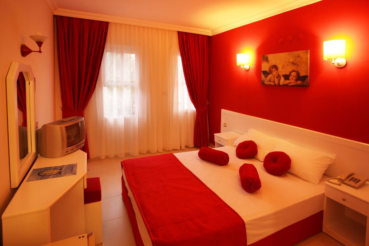 A hotel room with a large bed, red bedding, a television, a bedside table, and a painting on the wall.