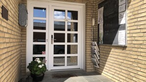 Un bâtiment en briques avec une porte blanche à deux battants, une petite plante en pot et un tableau noir affichant des informations.
