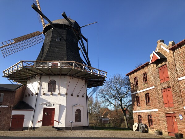 A traditional windmill with a white base and a black tower, adjacent to a brick building with red shutters and a sign indicating the year 1857. - Motel Apartments Tønder (Tonder)