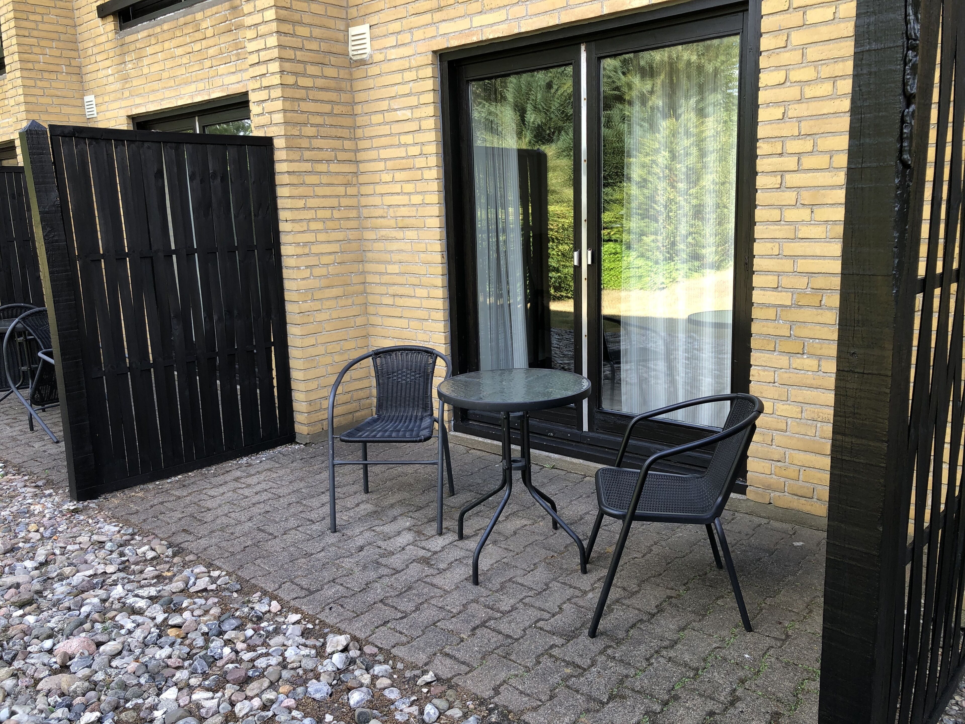 An outdoor patio with two metal chairs and a small round table, a black metal fence, and a yellow brick building with large glass doors.