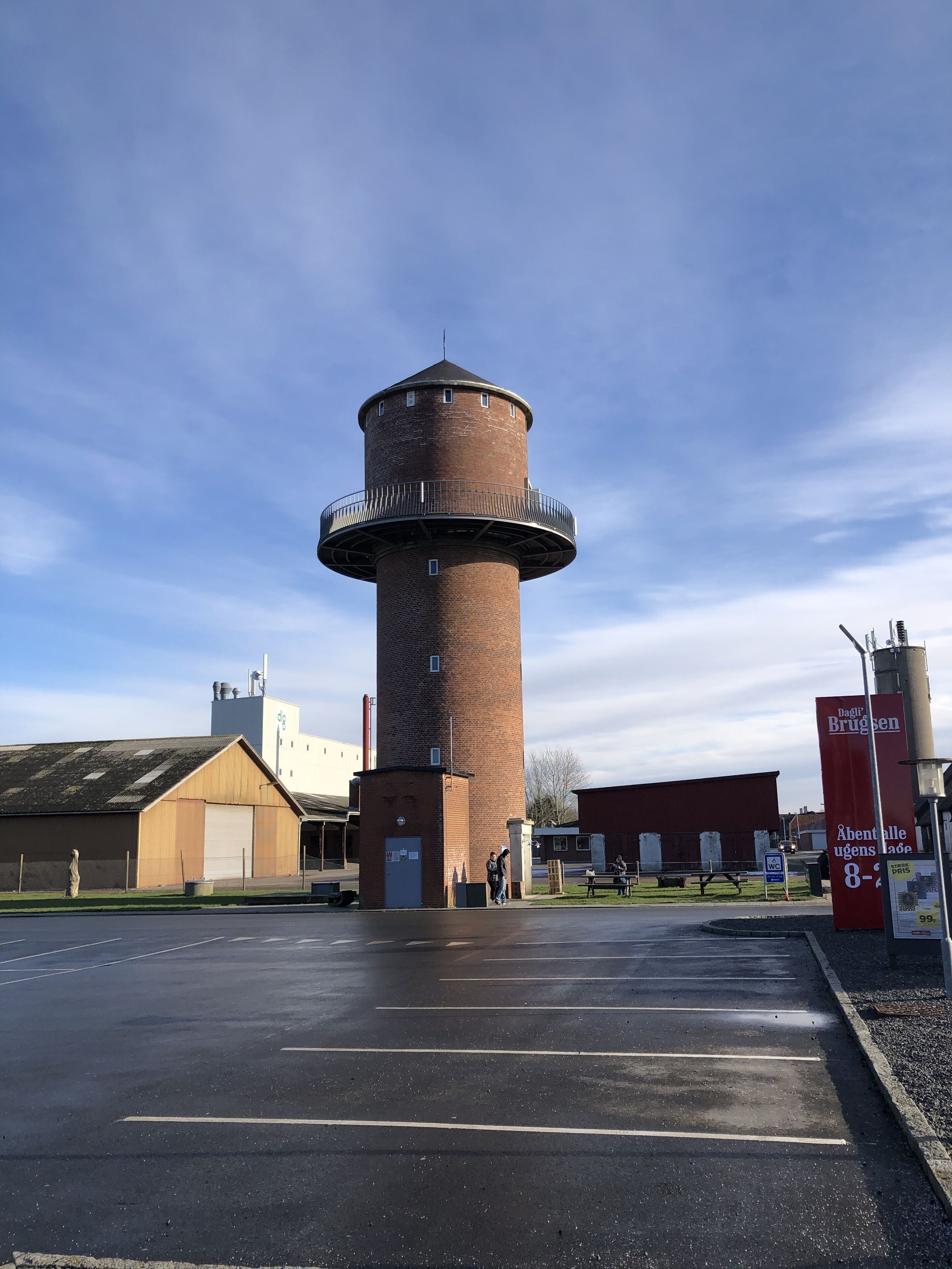 A brick lighthouse with a circular base and a balcony, surrounded by a parking lot and buildings.
