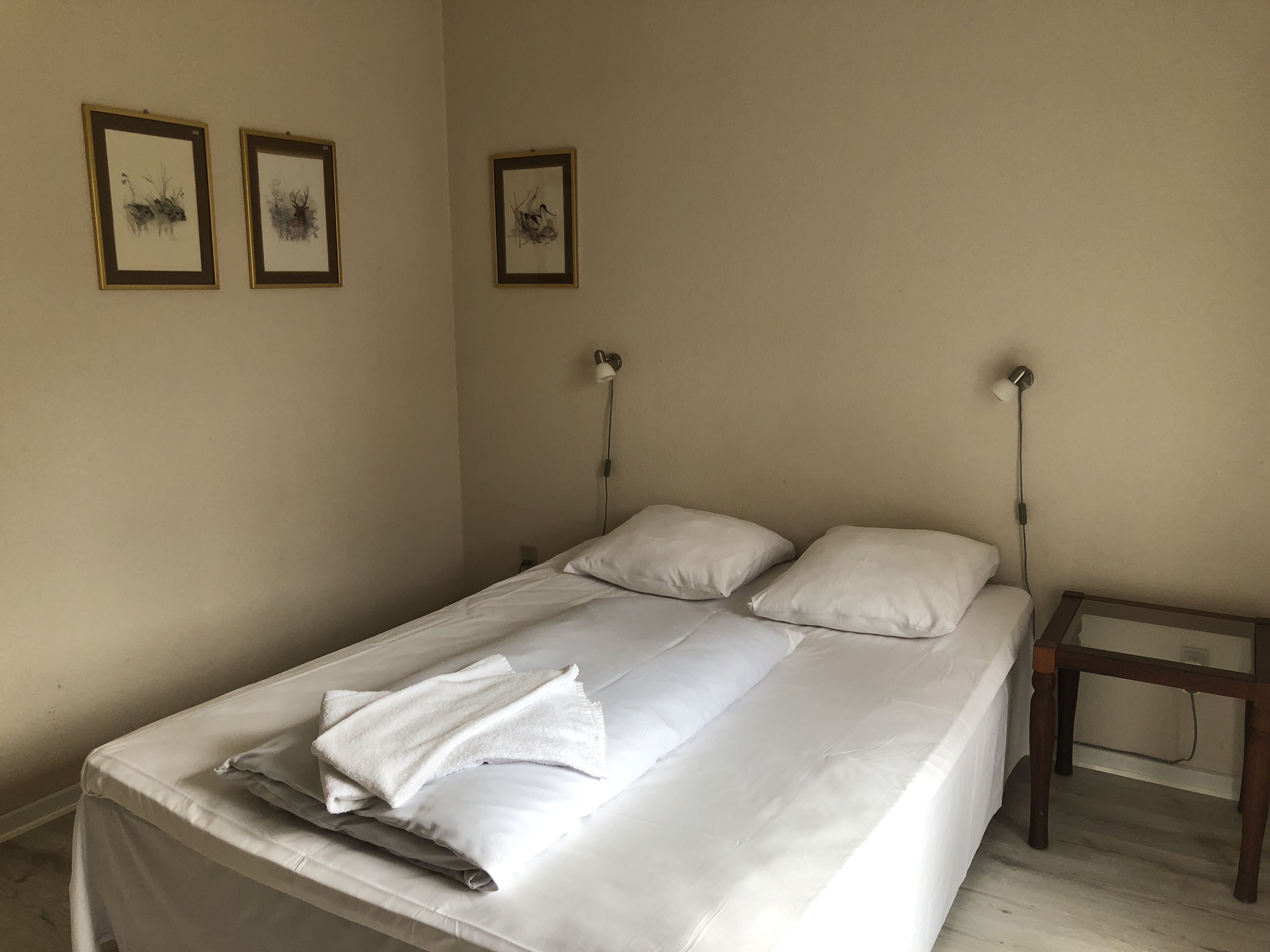 A neatly made bed with white linens and pillows, a wooden nightstand with a lamp, and three framed artworks on the wall.