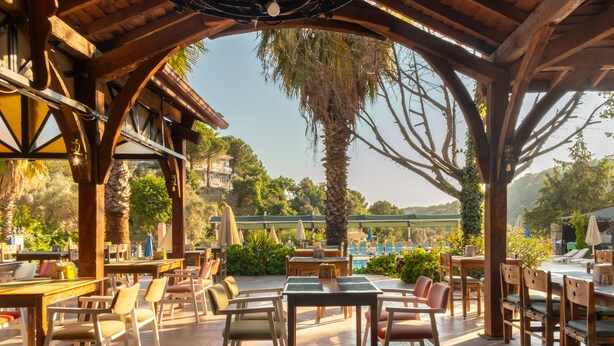 An outdoor patio area with wooden tables and chairs, a pool in the background, and a view of the surrounding landscape. - Pink Palace Hotel (Fethiye)