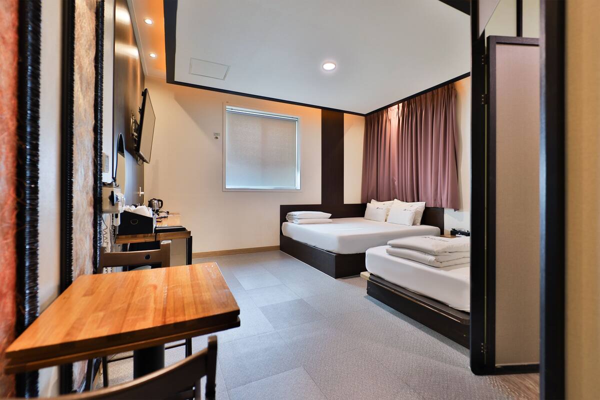 A modern hotel room with a wooden table, two beds, a desk, and a large window.