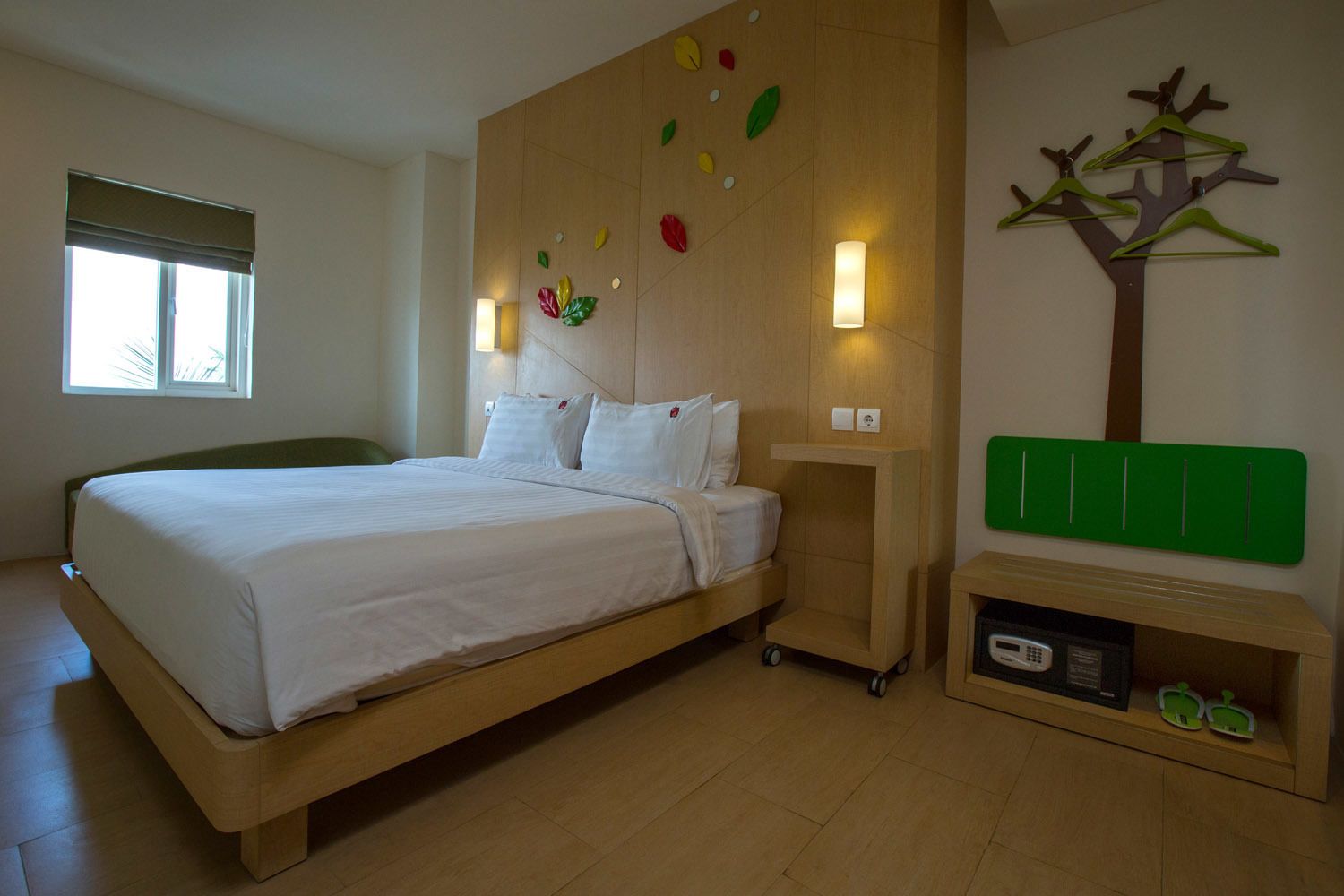 A bedroom with a bed, a nightstand, a wall-mounted tree decoration, and a small shelf with a microwave and a green object.