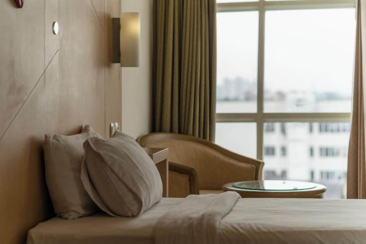 A hotel room with a bed, a chair, a small table, and a view of a cityscape through a window.