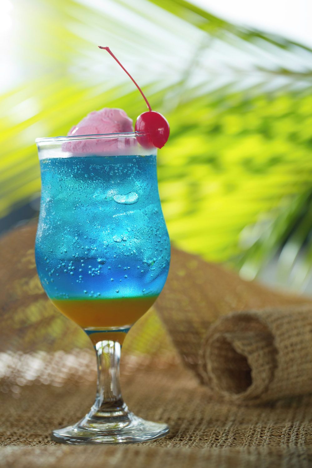 A cocktail glass with a blue liquid, a cherry, and a pink ice cube.
