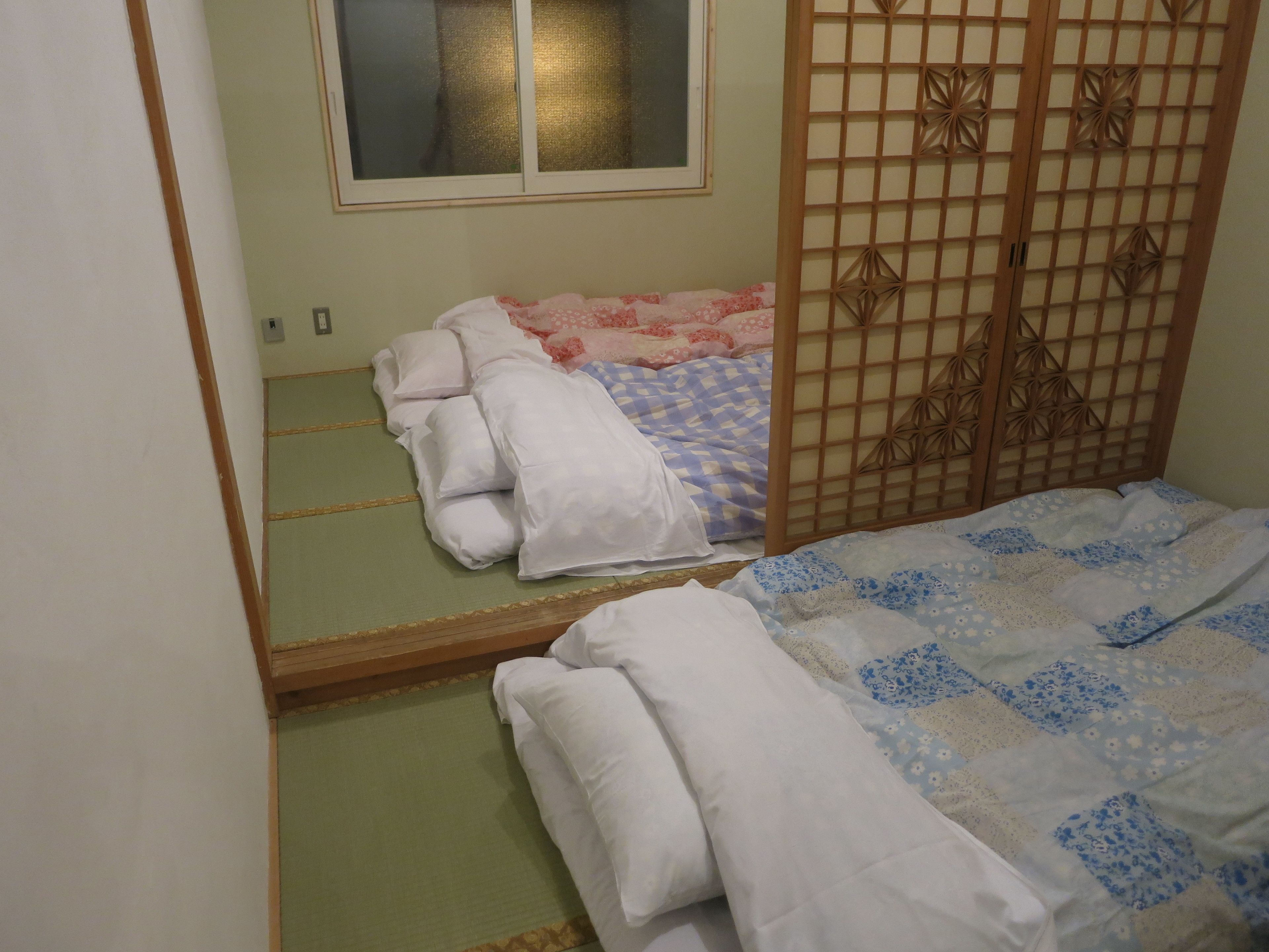 Ein traditioneller japanischer Raum mit Tatami-Boden, einem niedrigen Bett mit gemustertem Überwurf und einer Schiebetür mit geometrischen Mustern.