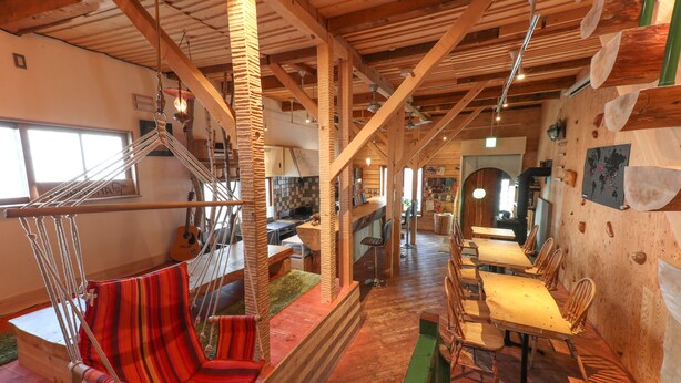 A spacious room with a wooden ceiling, a hammock chair, a dining area with wooden tables and chairs, and a kitchen in the background. - SappoLodge - Hostel (Sapporo)