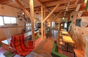 A spacious room with a wooden ceiling, a hammock chair, a dining area with wooden tables and chairs, and a kitchen in the background. - SappoLodge - Hostel (Sapporo)
