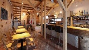A restaurant with wooden tables and chairs, a bar area with bottles, and a staircase leading to an upper level. - SappoLodge - Hostel (Sapporo)