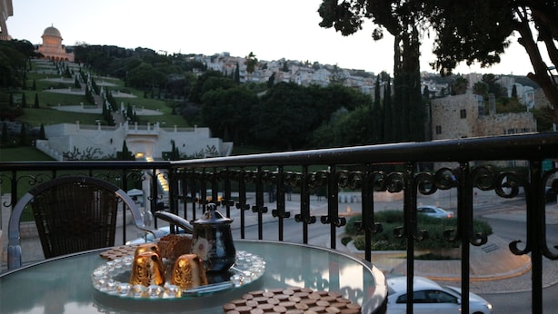 A balcony with a glass table, a wicker chair, and a view of a hill with buildings and trees. - Santa Maria Guest House (Haifa)