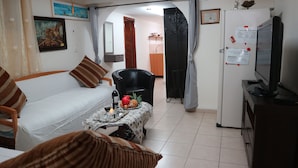 A living room with a sofa, a dining table with fruit and wine, a TV, and a refrigerator. - Santa Maria Guest House (Haifa)