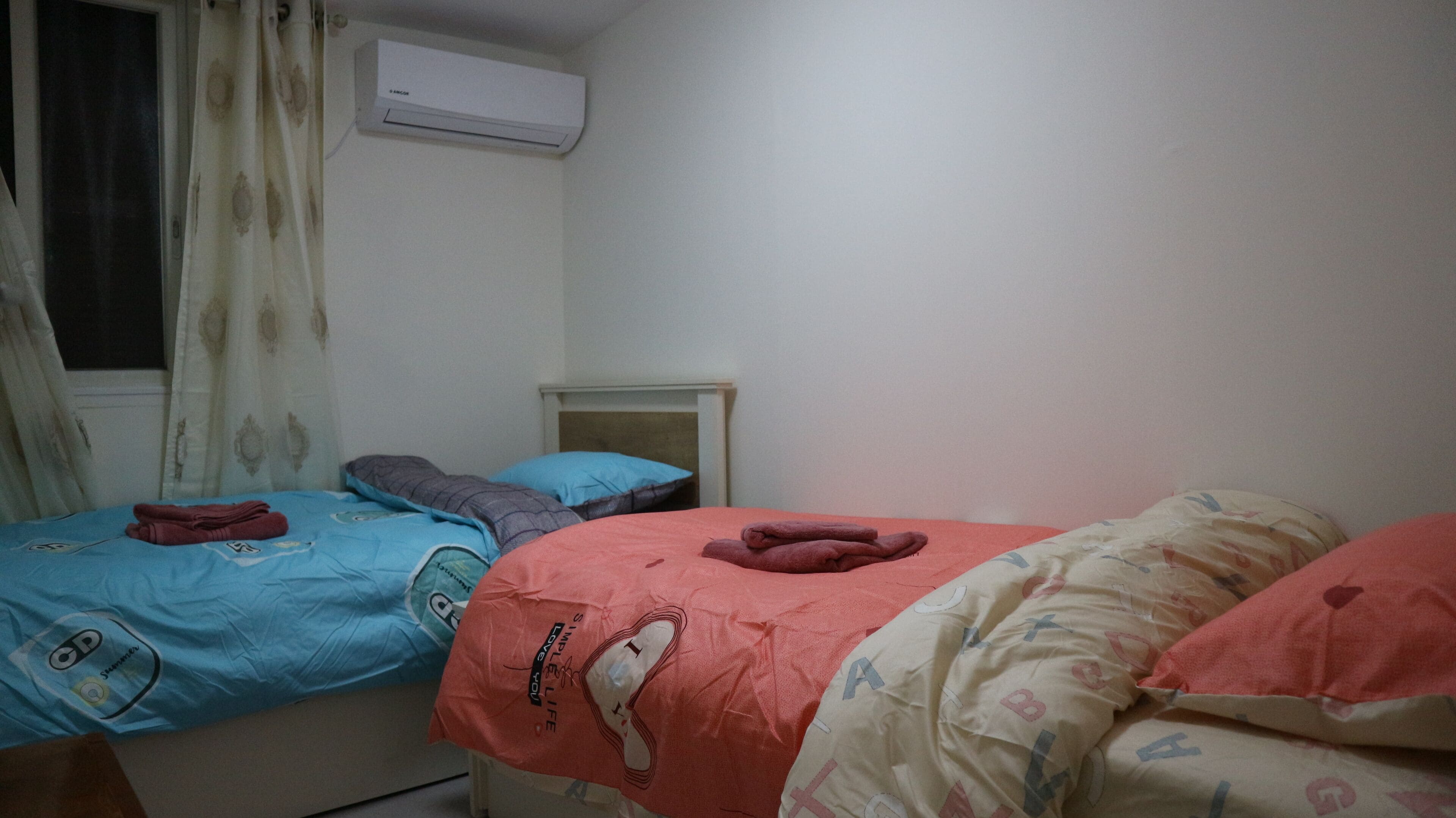 Een slaapkamer met twee bedden, één met een blauw dekbed en de ander met een rood dekbed. Er is een raam met gordijnen, een nachtkastje en een airconditioner aan de muur.