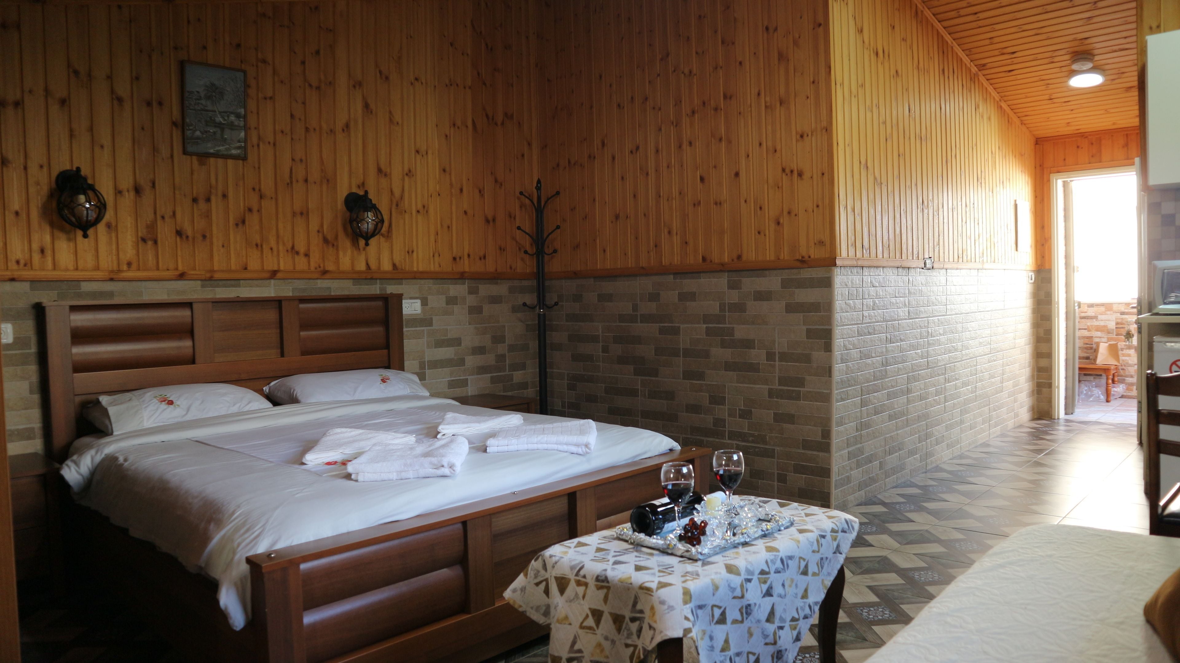 Una habitación con paneles de madera, una cama, una mesa con una copa de vino y un perchero.