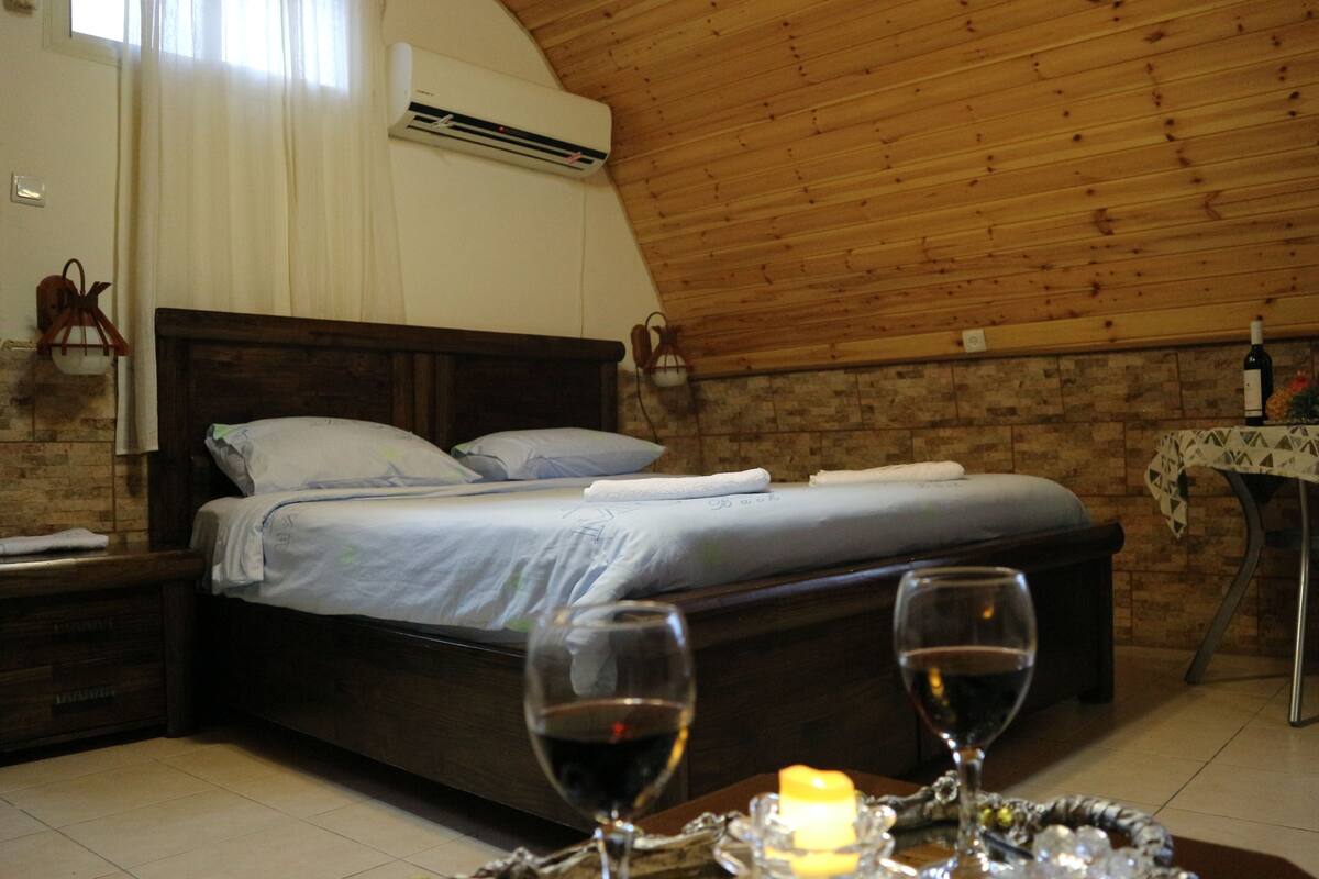 A cozy bedroom with a wooden ceiling, a bed with white linens, a bedside table with a wine glass, and a small table with a candle and decorative items.