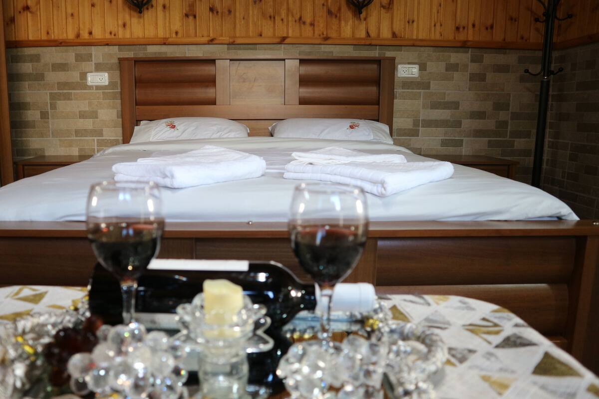 A neatly made bed with white linens and towels, two wine glasses on a table with a candle and decorative items.