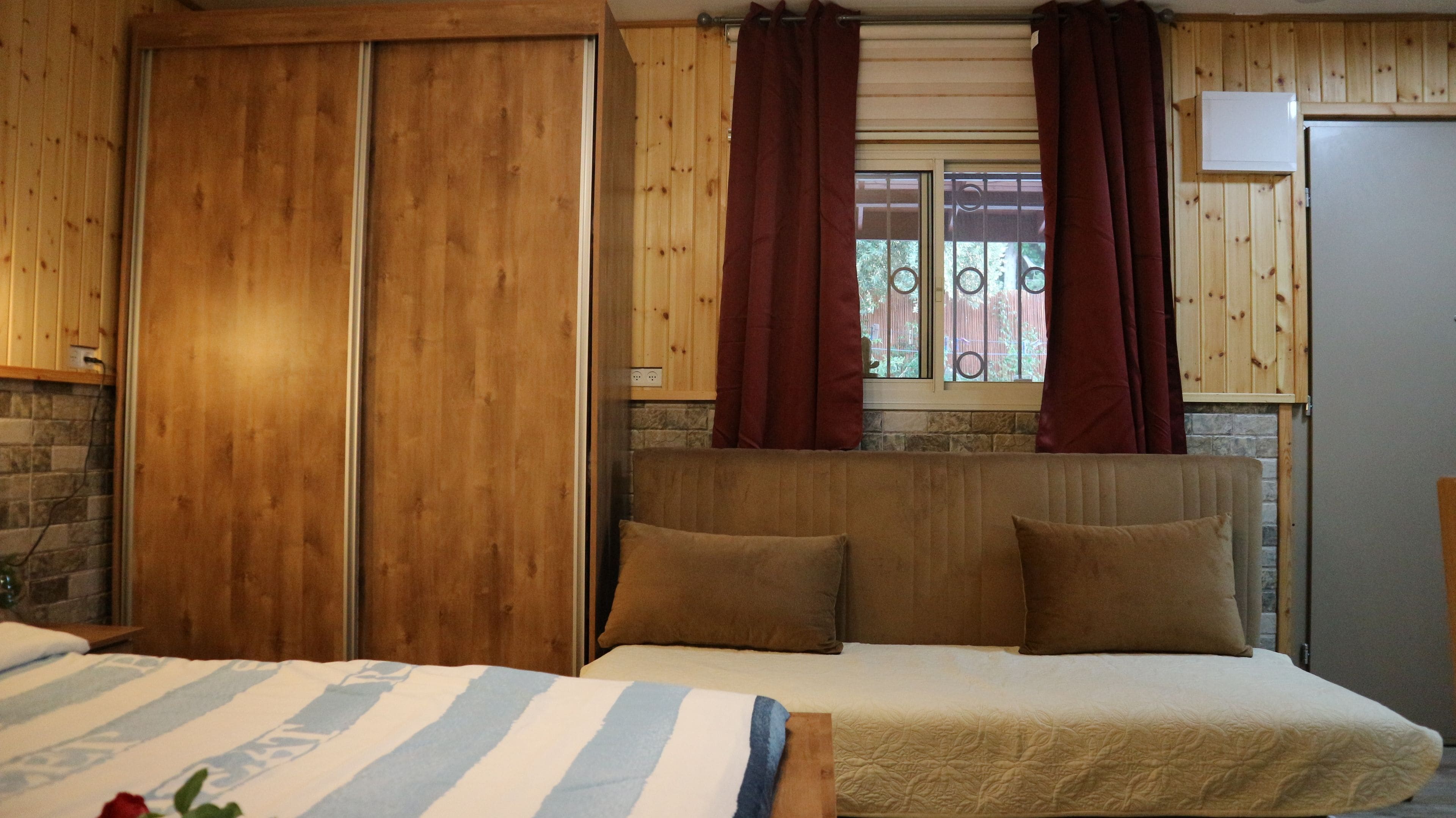 Een slaapkamer met een houten hoofdeinde, twee kussens, een gestreept dekbed en een raam met gordijnen.