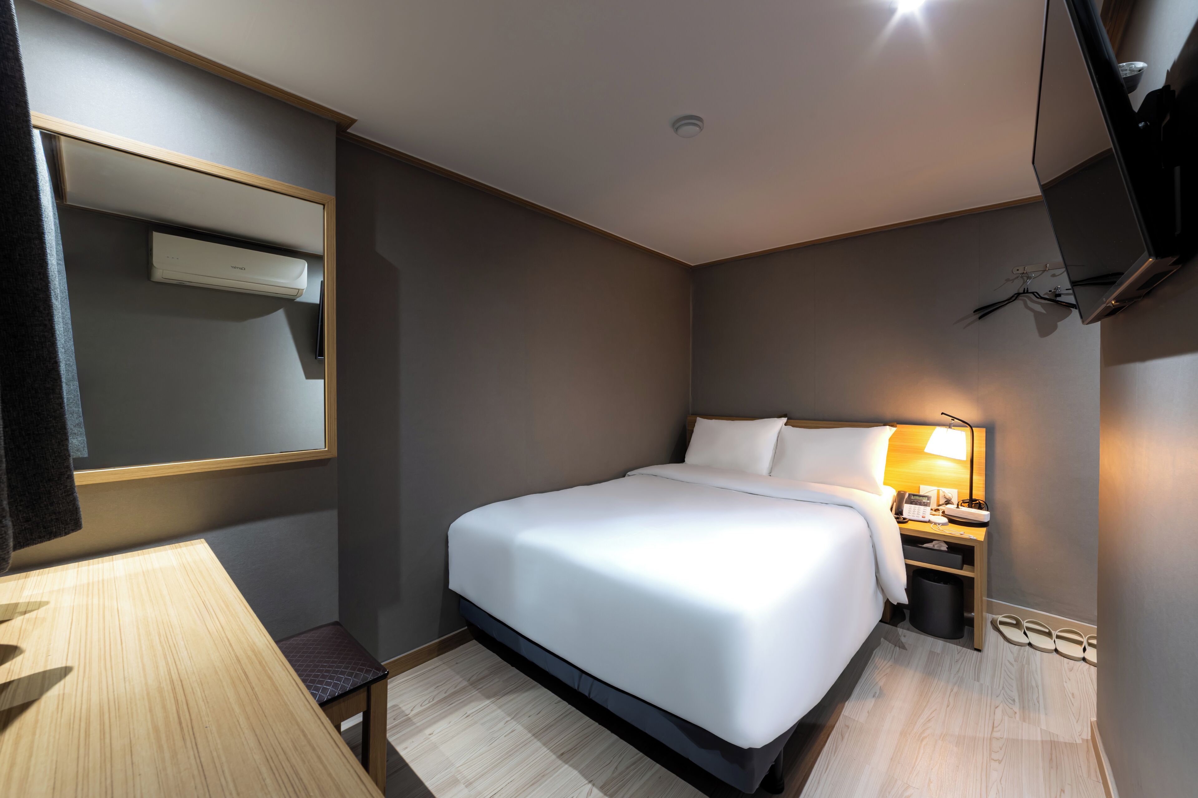 A hotel room with a bed, bedside table, lamp, and a TV mounted on the wall.