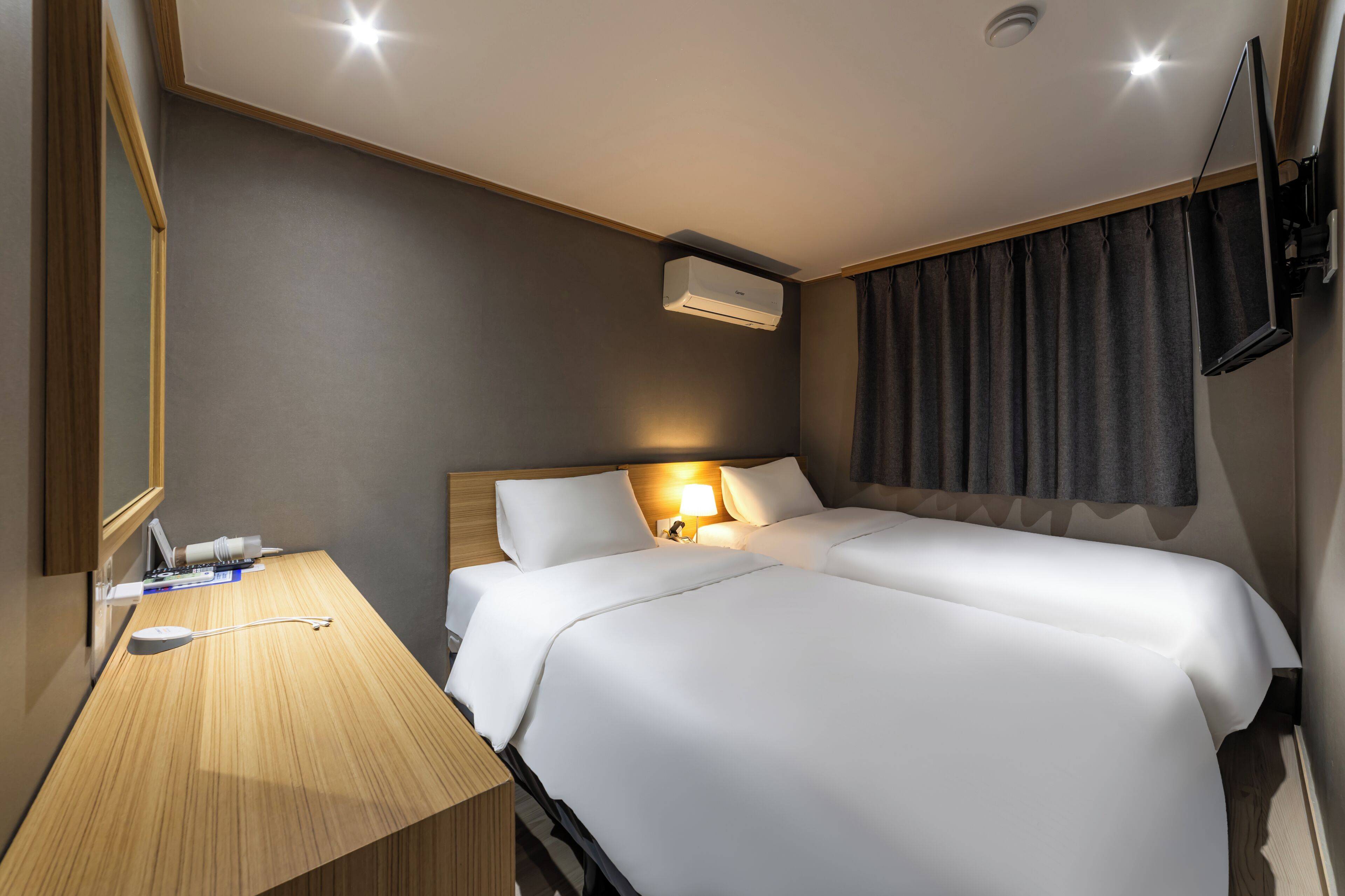 A hotel room with two beds, a desk, a flat-screen TV, and an air conditioning unit.