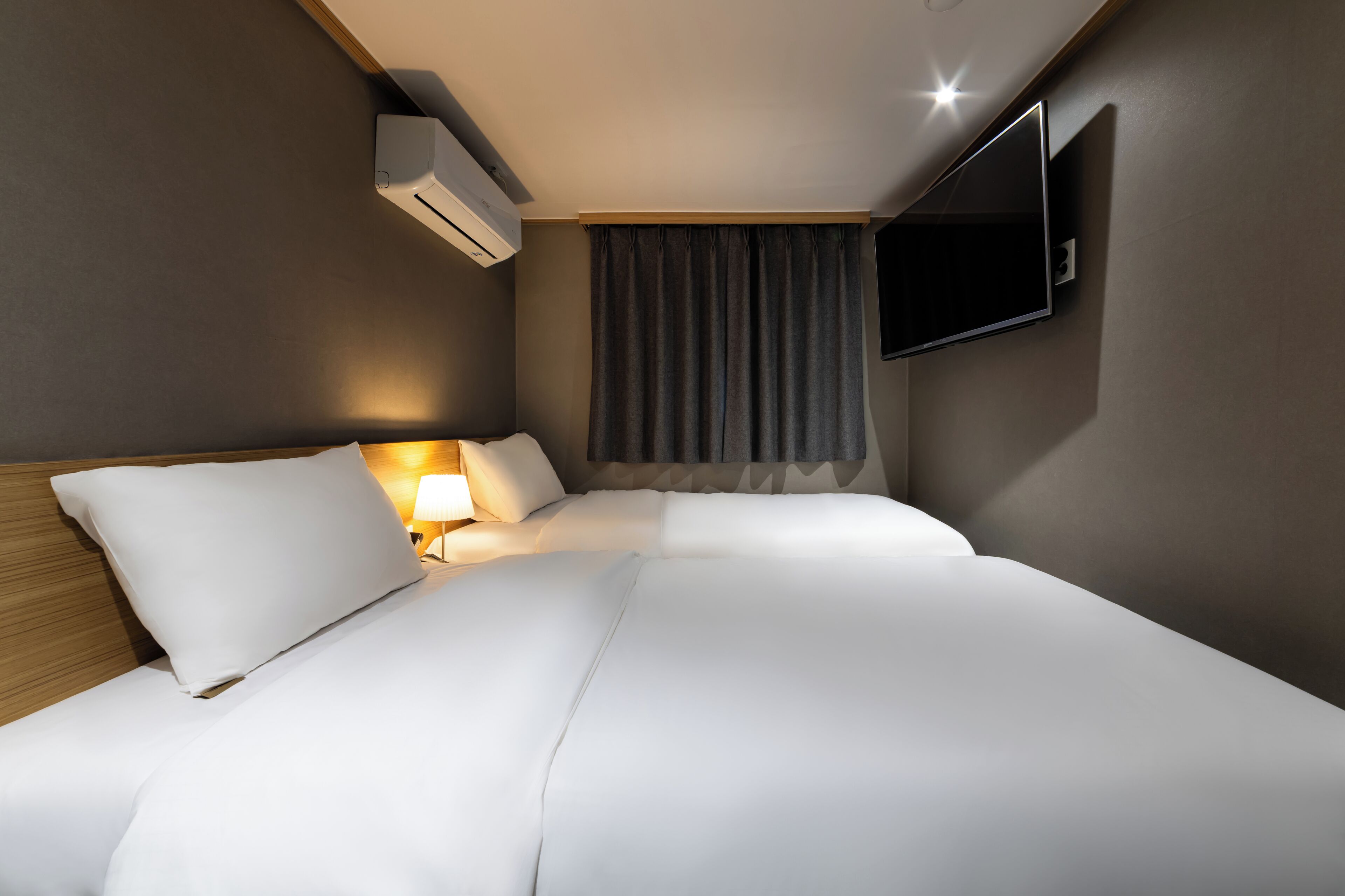 A hotel room with a bed, a TV mounted on the wall, an air conditioning unit, and a window with curtains.
