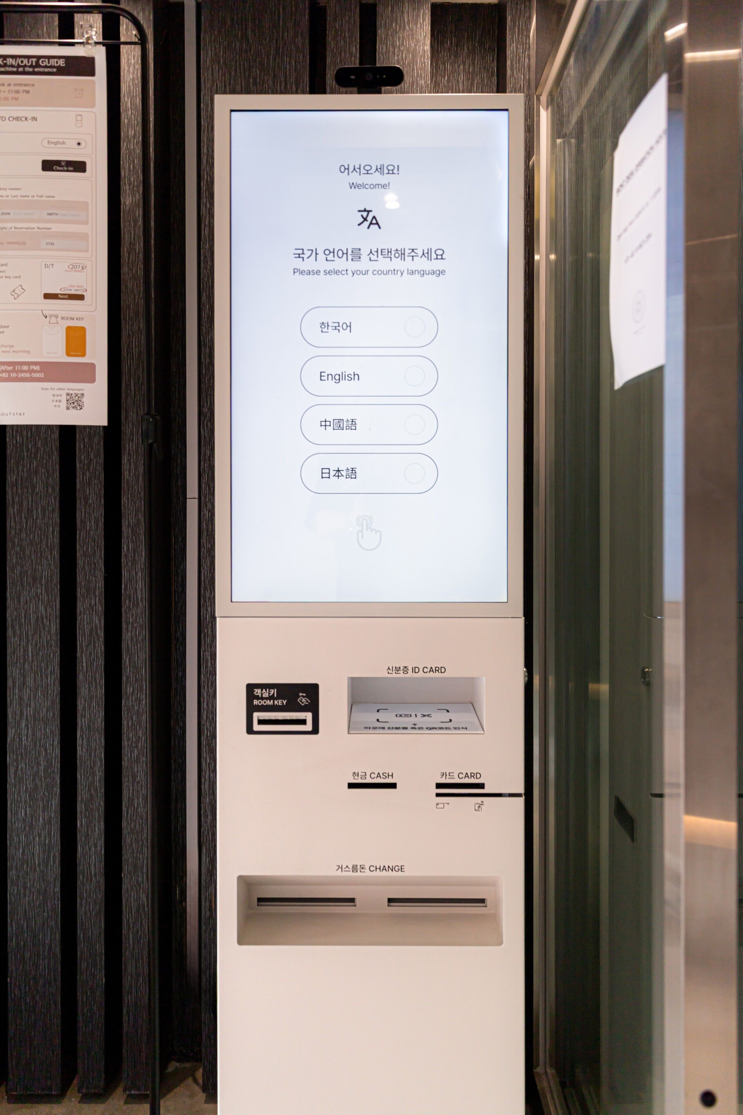 Een digitale kiosk met een scherm waarop een taalmenu, een sleuf voor een hotelpasje en een kaartlezer voor contante en kaartbetalingen staan.