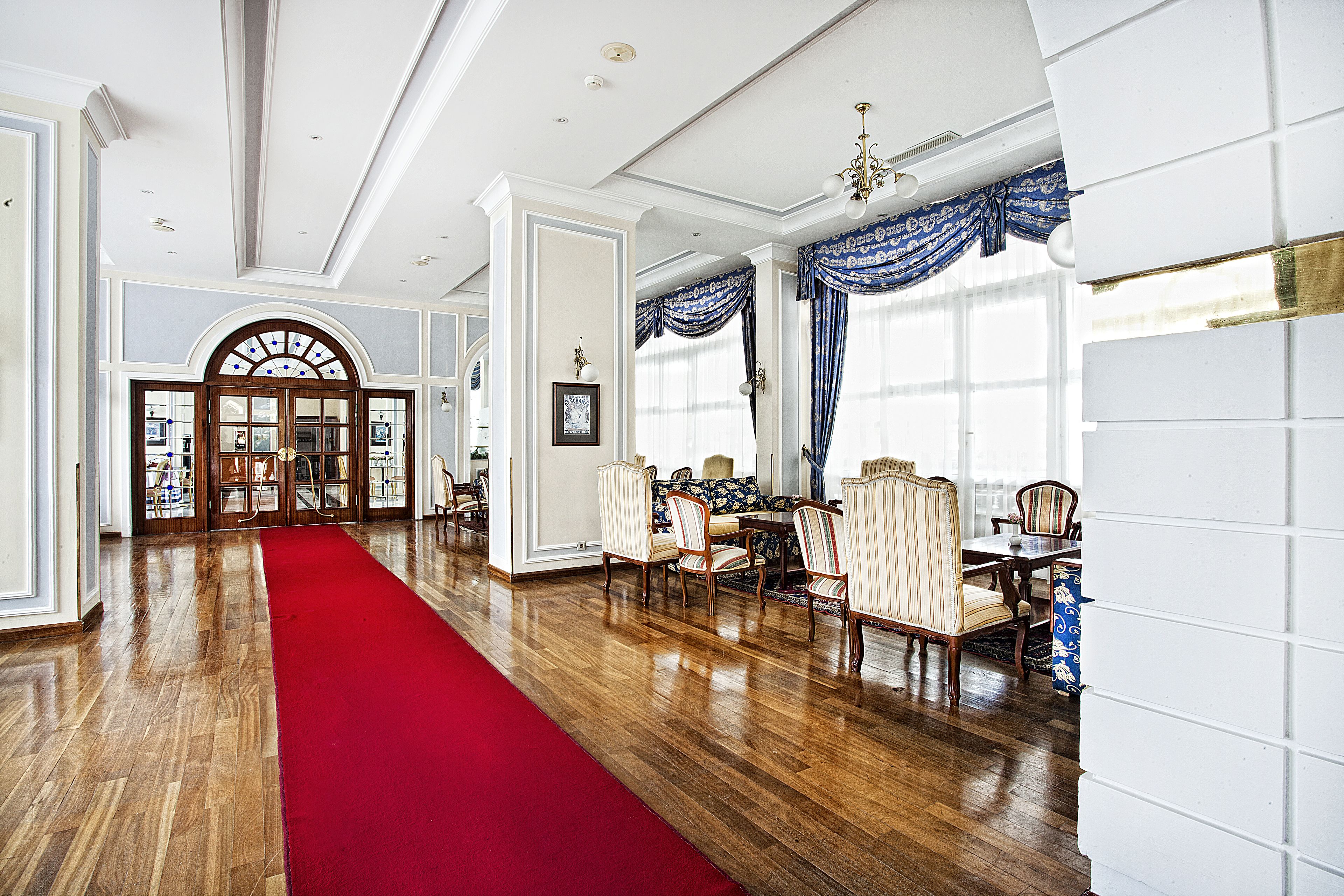 A spacious dining area with a red carpet leading to a large window, wooden flooring, and classic furniture.