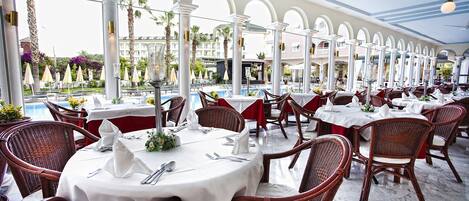 A restaurant with round tables, wicker chairs, and a view of a pool and palm trees.