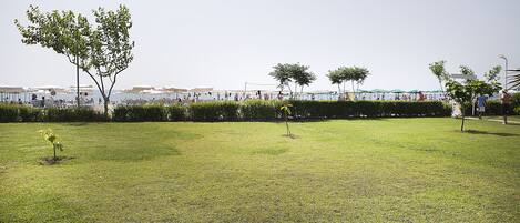 Un espace extérieur verdoyant, avec des arbres, d’où l’on peut apercevoir une plage et, au loin, des parasols.