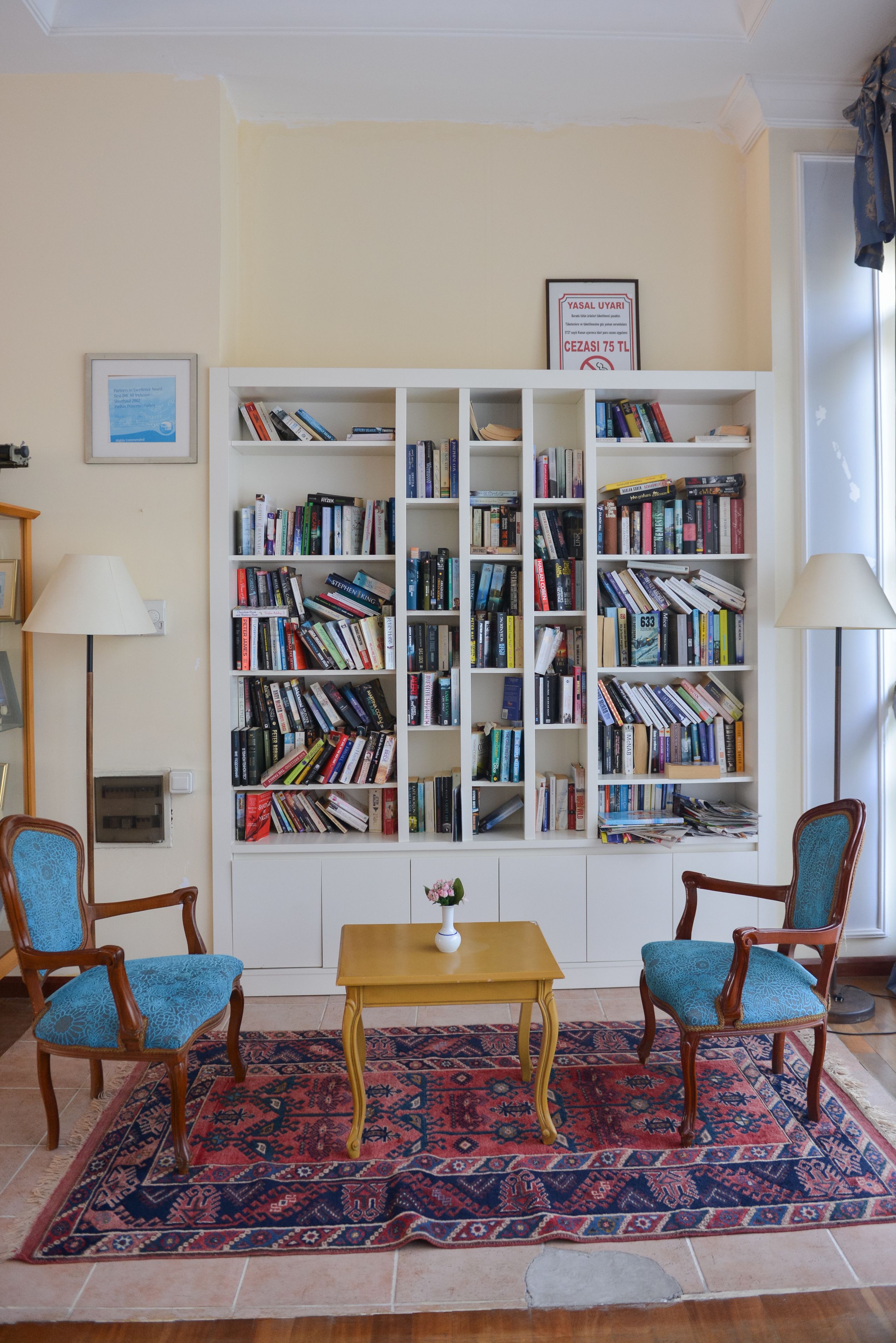 A living room with a large bookshelf, two blue upholstered chairs, a small wooden table, and a patterned rug.
