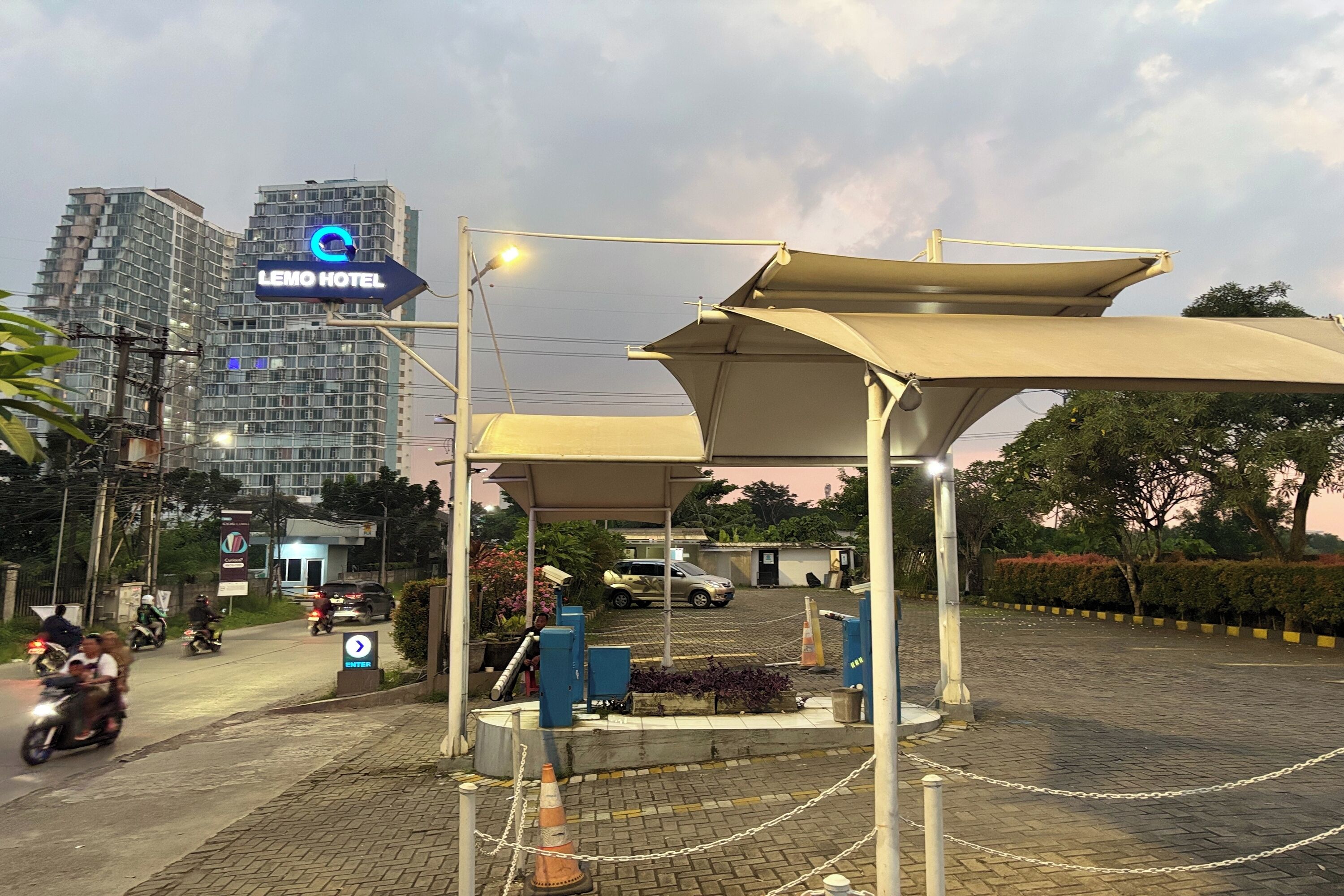 Entrada de um hotel com área de espera coberta, placa indicando LEMO HOTEL e estacionamento com veículos.