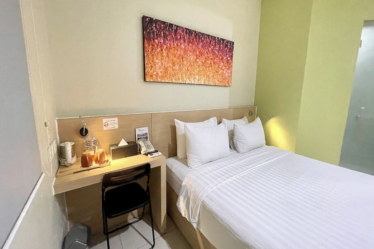 A hotel room with a bed, a desk with a telephone, a chair, and a small table with cups and a coffee maker.