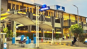 Entrada de hotel con una zona de espera cubierta, estacionamiento para coches y una cancela visible.