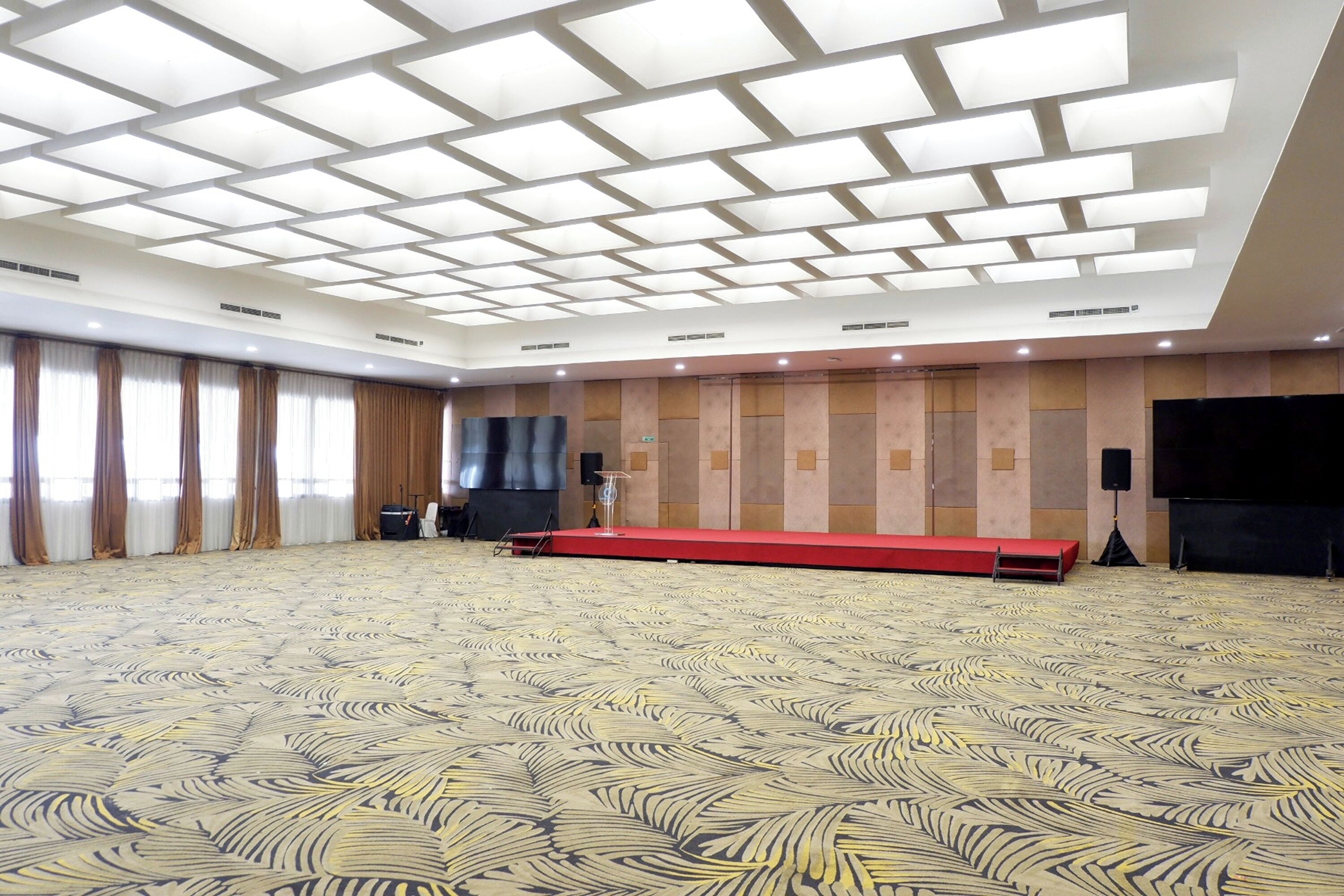 A large conference room with a patterned carpet, a stage with a red platform, and a large flat-screen TV on the wall.