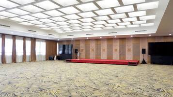 A large conference room with a patterned carpet, a stage with a red platform, and a large flat-screen TV on the wall.