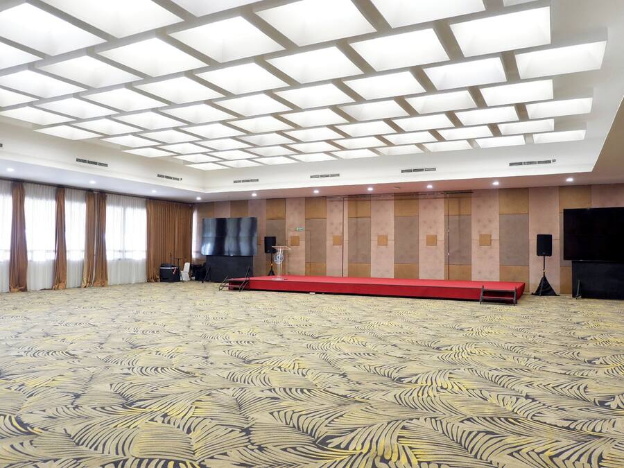 A large conference room with a patterned carpet, a stage with a red platform, and a large flat-screen TV on the wall.