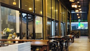 Un restaurante con mesas y sillas largas, ventanas amplias y lámparas colgantes.
