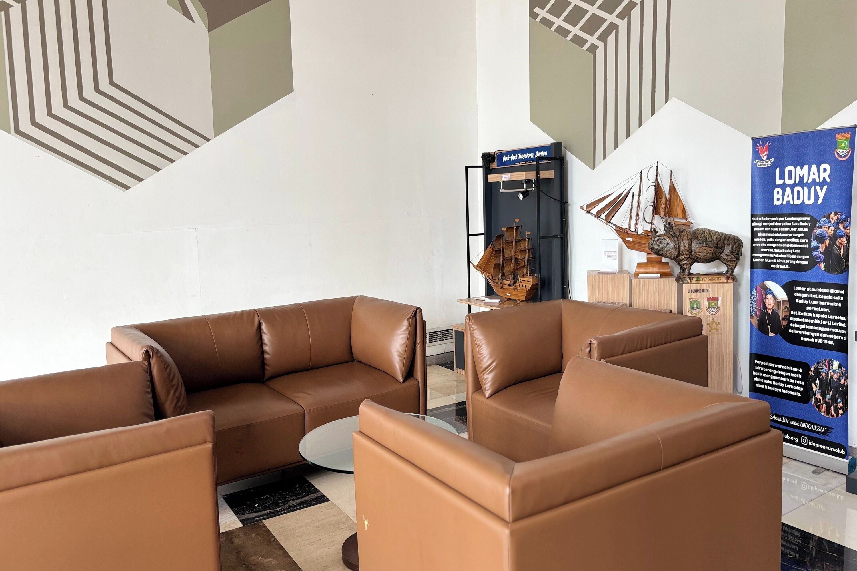 A modern lobby with brown leather sofas, a glass coffee table, and a decorative sailboat. A signboard with 'LOMAR BADUY' is visible.