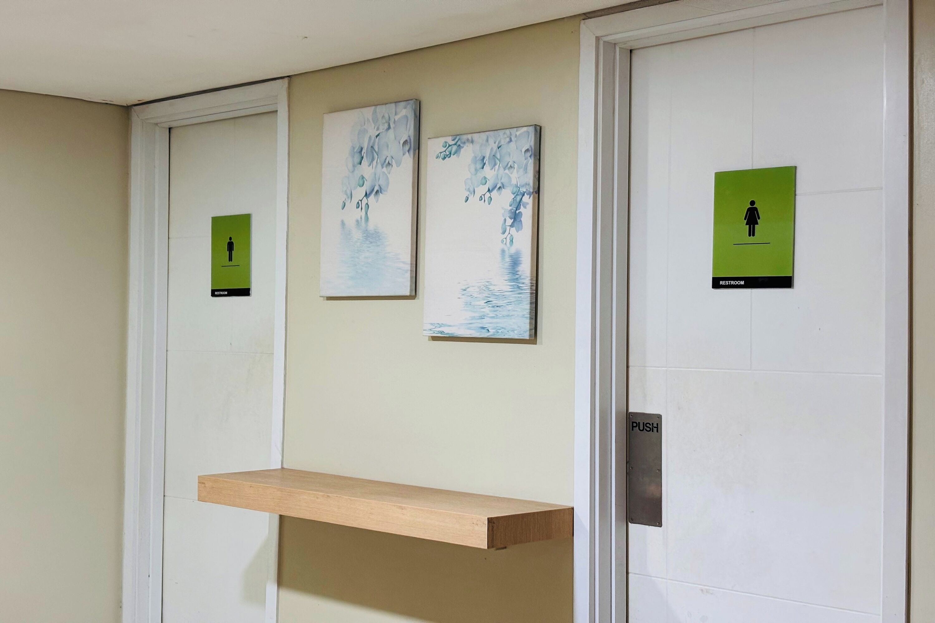 A restroom with a push button, a wooden shelf, and three signs indicating restrooms and a women's restroom.