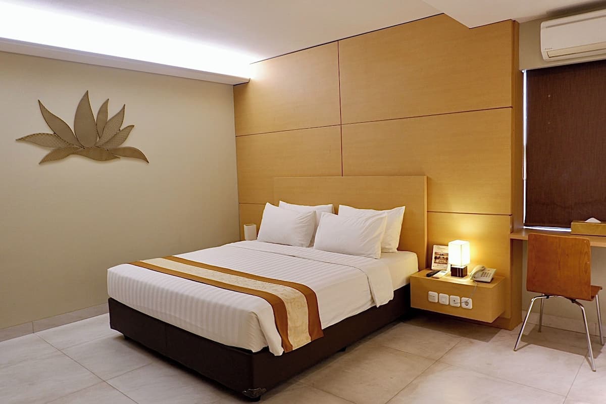 A hotel room with a large bed, a bedside table with a lamp, a chair, and a wall-mounted air conditioner.