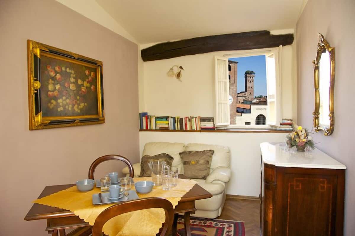 A dining area with a table set for two, a bookshelf, a window with a view of a tower, and a mirror on the wall.