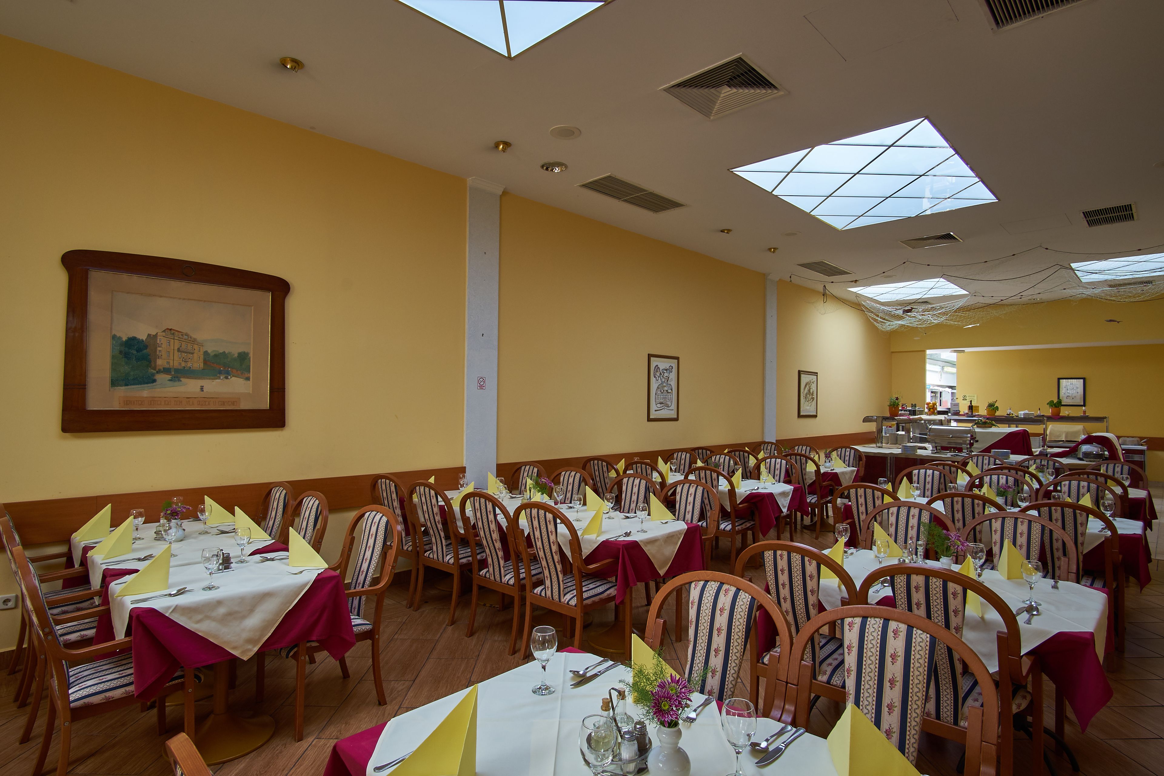 A large dining hall with multiple tables set for dining, wooden chairs, and yellow tablecloths.