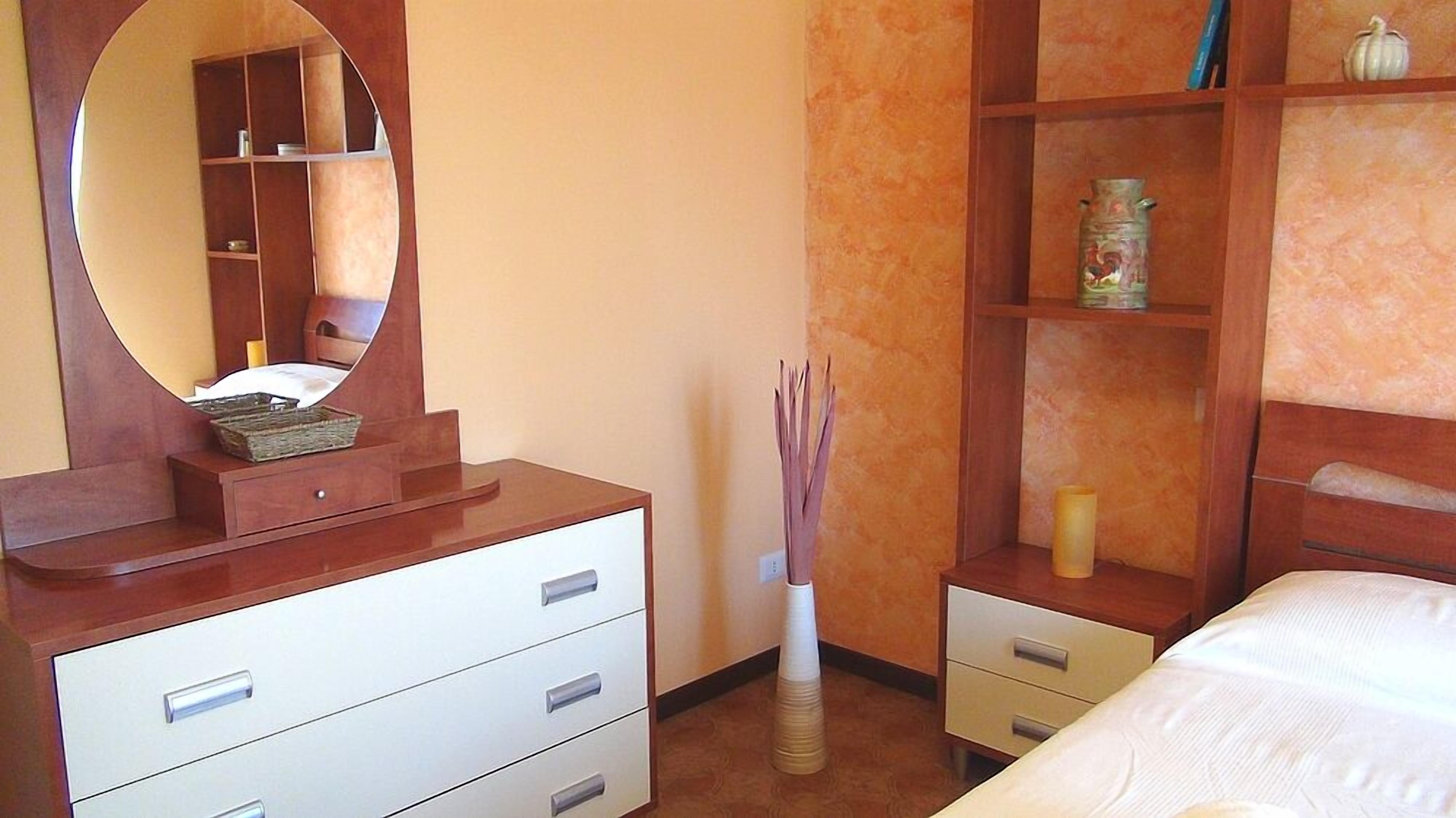 A bedroom with a wooden dresser, a mirror, a bedside table, a bed, and a shelf with decorative items.