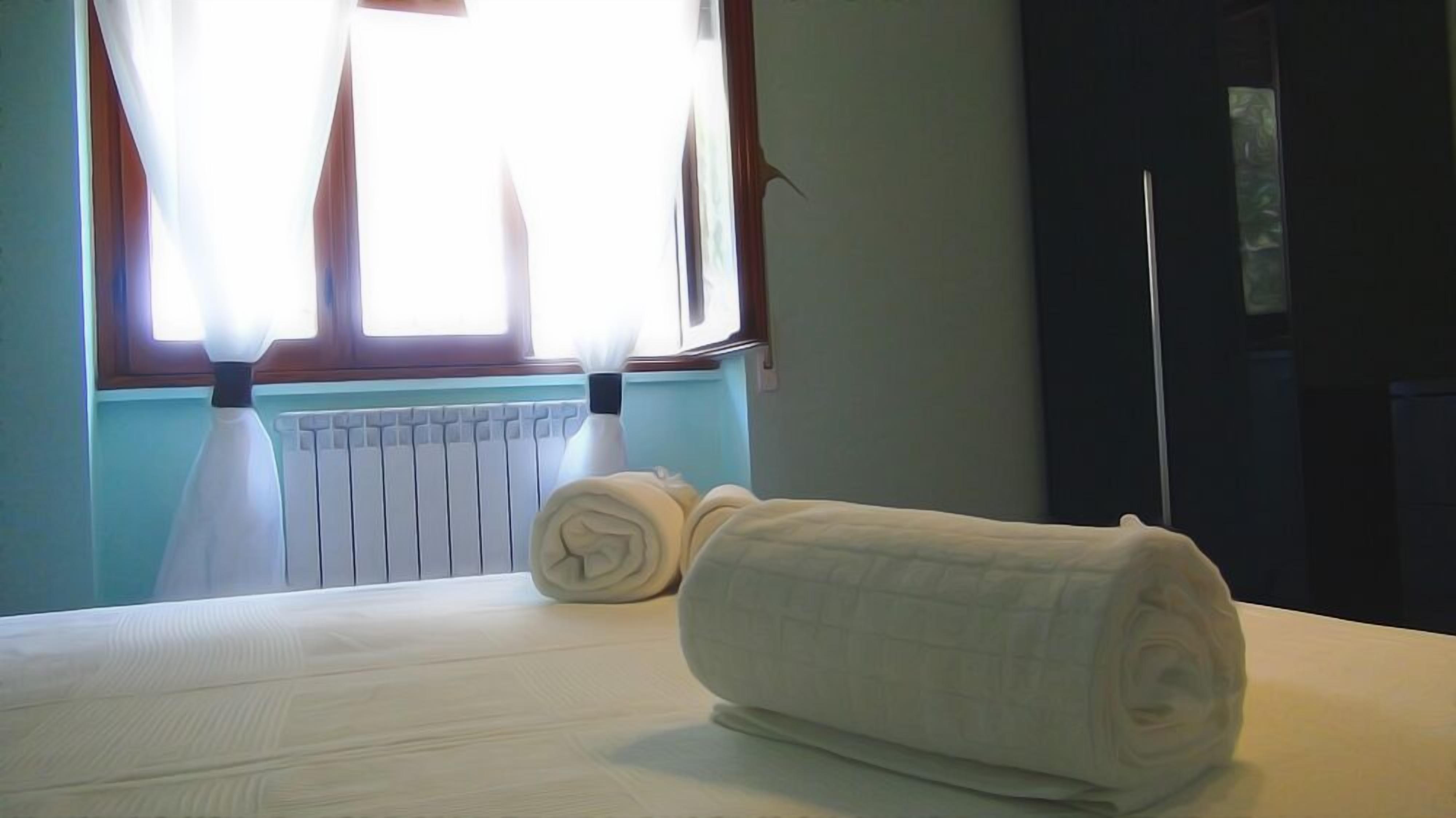 A hotel room with a bed, a rolled towel, a radiator, and a window with curtains.