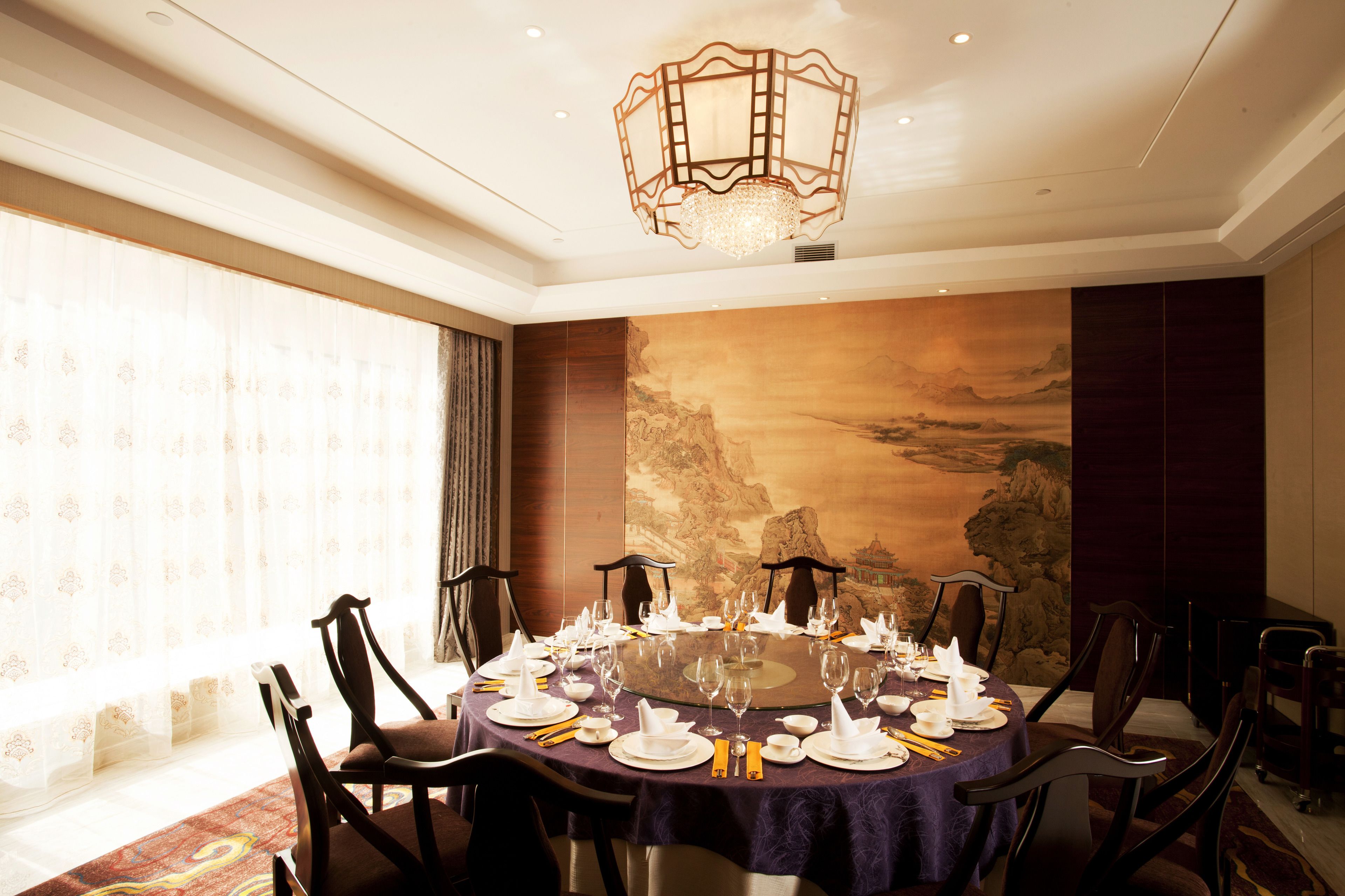 Mesa de jantar redonda posta para uma refeição, com cadeiras ao redor, um lustre acima e um mural grande na parede.