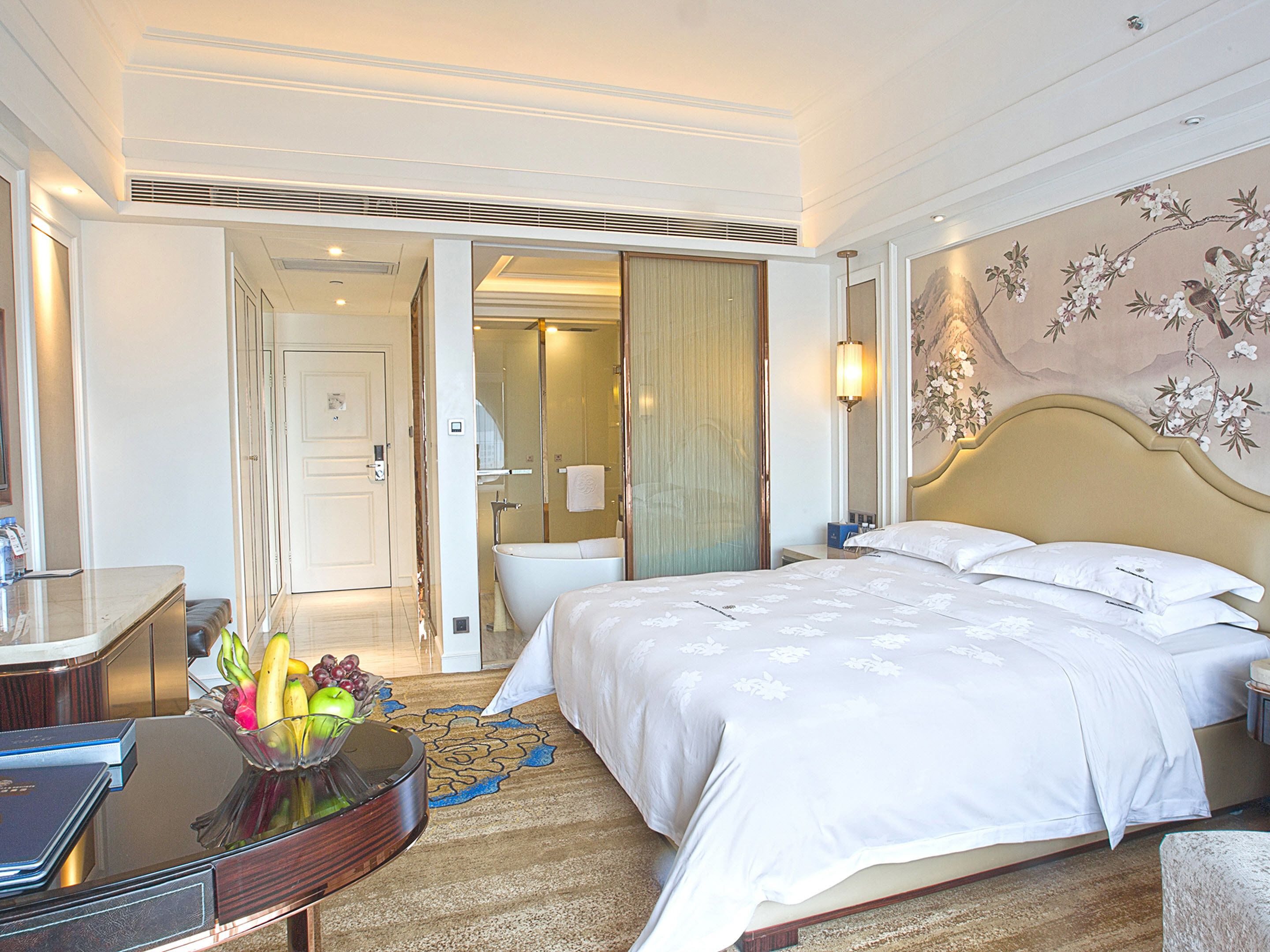 Habitación de hotel con una cama grande, un escritorio con fruteros, un baño con bañera y un adorno de pared.