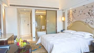Habitación de hotel con una cama grande, un escritorio con fruteros, un baño con bañera y un adorno de pared.
