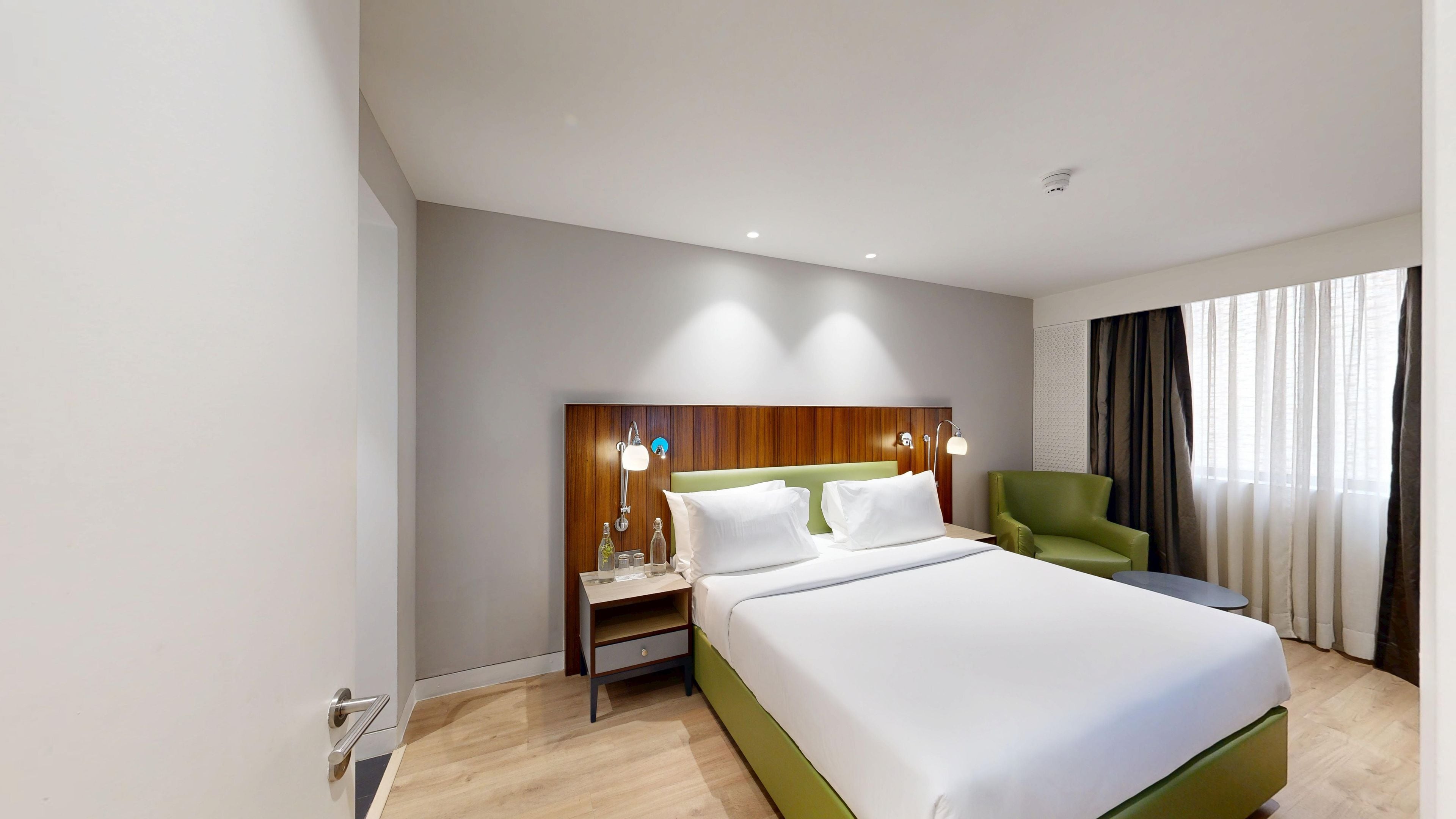 A hotel room with a bed, a nightstand, a green chair, and a window with curtains.
